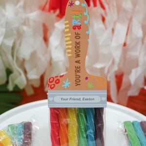 May include: A paintbrush-shaped gift with rainbow-colored candy 'bristles'. The handle is tan with the text 'YOU'RE A WORK OF ART!' and 'Your Friend, Easton'. A creative and colorful treat, perfect for art lovers.
