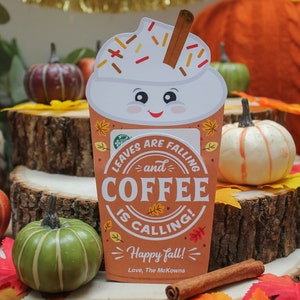 Może przedstawiać: Festive holder na kartę podarunkową w kształcie kubka z kawą, z bitą śmietaną i cynamonową pałeczką. Na holderze widnieje tekst "Leaves are falling and coffee is calling! Happy fall! Love, The McKowns".