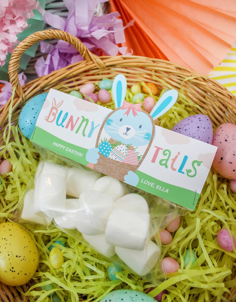 May include: A wicker basket filled with pastel-colored shredded paper and candy eggs. A clear plastic bag with white bunny tail marshmallows is in the basket. A colorful tag with a bunny illustration and the text "Bunny Tails" is attached to the bag. The tag also says "Happy Easter! Love, Ella."