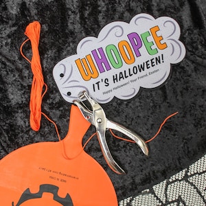 May include: A close-up of a Halloween craft project. A white card with the text "WHOOPEE IT'S HALLOWEEN! Happy Halloween! Your Friend, Easton" is attached to an orange pumpkin-shaped balloon with a hole punched in it. A pair of silver hole punch pliers and orange string are also visible.