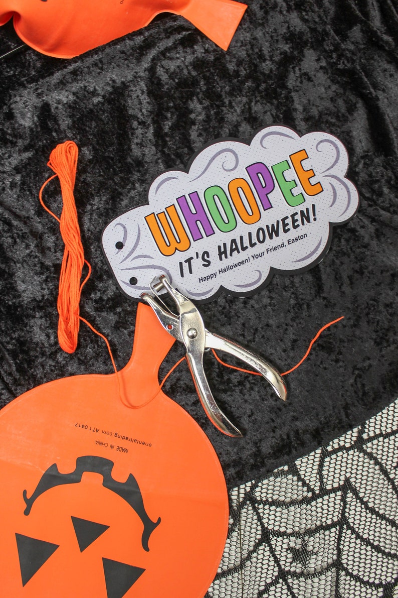 May include: A close-up of a Halloween craft project. A white card with the text "WHOOPEE IT'S HALLOWEEN! Happy Halloween! Your Friend, Easton" is attached to an orange pumpkin-shaped balloon with a hole punched in it. A pair of silver hole punch pliers and orange string are also visible.