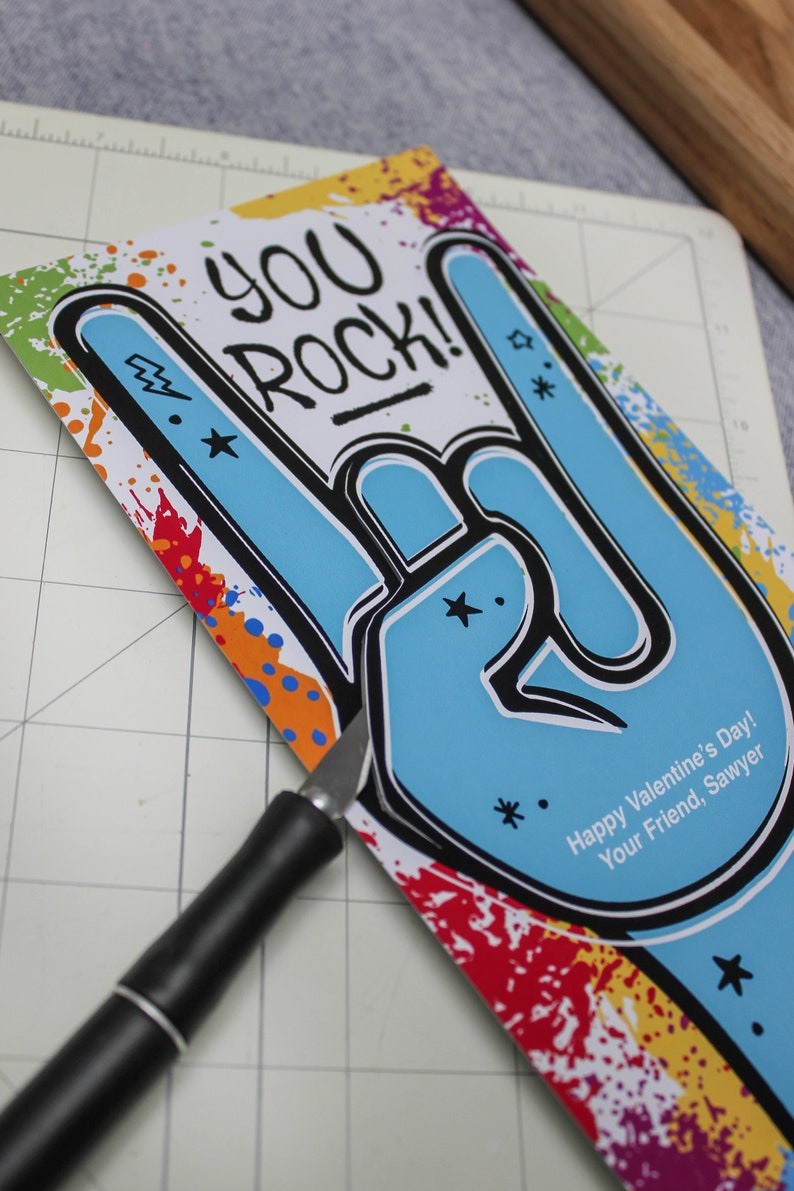 Pode incluir: Um cart&atilde;o colorido com uma m&atilde;o azul fazendo o gesto de rock and roll. O cart&atilde;o diz "You Rock!" e "Happy Valentine's Day! Your Friend, Sawyer".