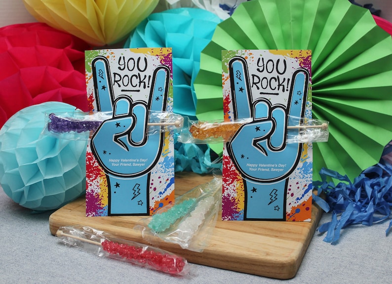 Pode incluir: Dois cart&otilde;es coloridos do Dia dos Namorados com o texto "You Rock!" e um gesto de m&atilde;o de rock and roll. Cada cart&atilde;o tem um pau de caramelo de rocha anexado.