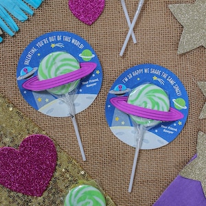 Op de afbeelding: Drie groene en witte swirl-lollipops met een ruimtethema. Elke lollipop is bevestigd aan een witte stok en heeft een ronde kaart met een roze ring, een ruimteschip en sterren. De tekst op de kaarten luidt "Valentine, je bent niet van deze wereld!" en "Ik ben zo blij dat we dezelfde ruimte delen!"