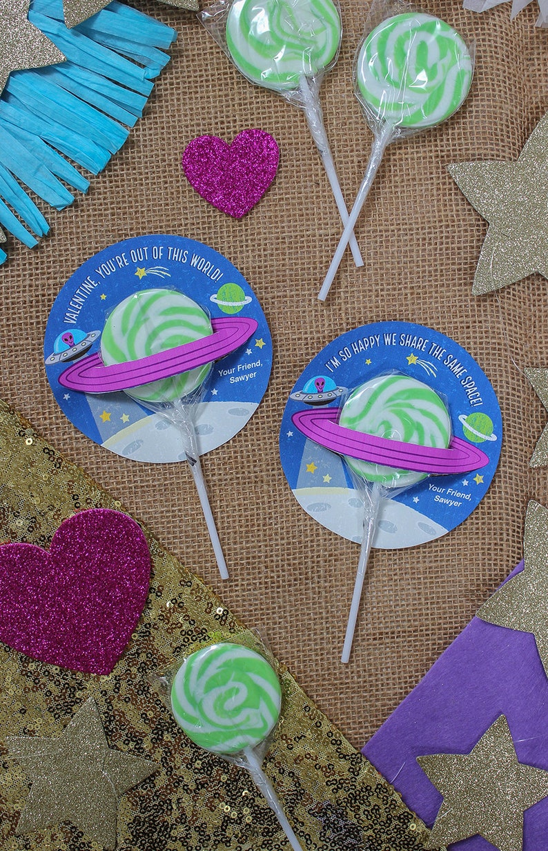 Op de afbeelding: Drie groene en witte swirl-lollipops met een ruimtethema. Elke lollipop is bevestigd aan een witte stok en heeft een ronde kaart met een roze ring, een ruimteschip en sterren. De tekst op de kaarten luidt "Valentine, je bent niet van deze wereld!" en "Ik ben zo blij dat we dezelfde ruimte delen!"