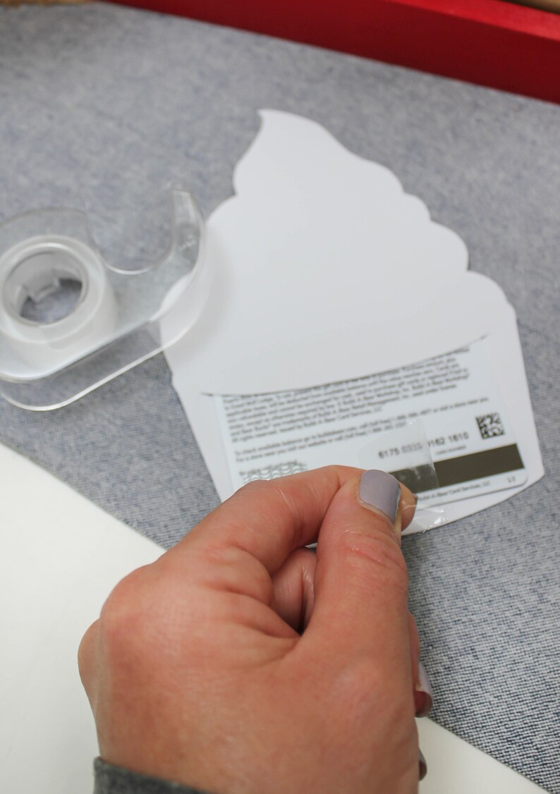 May include: A white cupcake-shaped gift card with a clear adhesive strip being peeled off. The gift card has a barcode and text on the back.