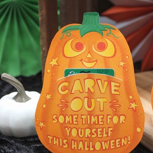 May include: Orange pumpkin-shaped card with the text "Carve Out Some Time For Yourself This Halloween!" and a green gift card tucked inside. The card has a smiling pumpkin face with yellow eyes and a white background. The text is in yellow and orange. The card is sitting on a black surface.