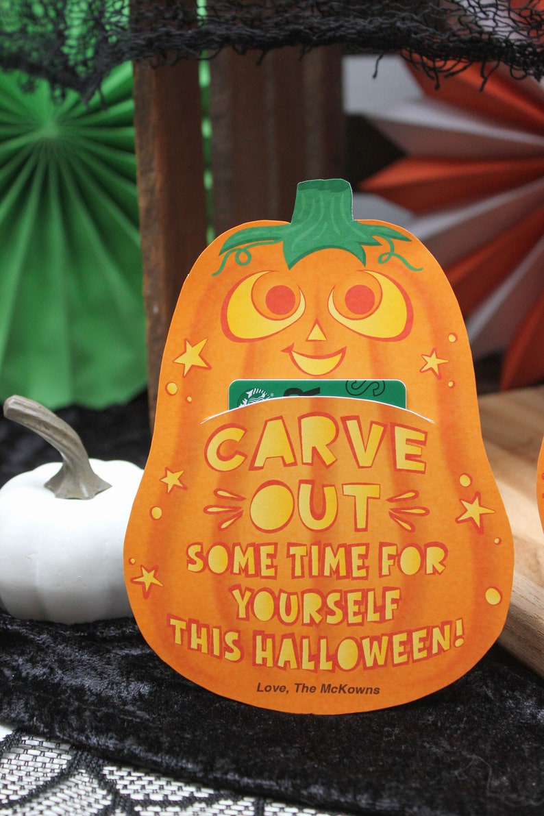 May include: Orange pumpkin-shaped card with the text "Carve Out Some Time For Yourself This Halloween!" and a green gift card tucked inside. The card has a smiling pumpkin face with yellow eyes and a white background. The text is in yellow and orange. The card is sitting on a black surface.