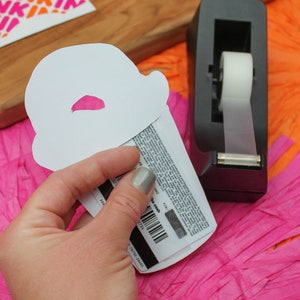 May include: A white paper cutout of a coffee cup with a pink lid. The cutout is being held over a Dunkin' Donuts gift card with a barcode and the text "Dunkin' Donuts Gift Card".