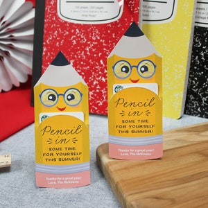 May include: Two yellow pencil-shaped gift card holders with smiling faces and glasses, with the text "Pencil in some time for yourself this summer!". The background includes composition notebooks and a decorative paper fan.