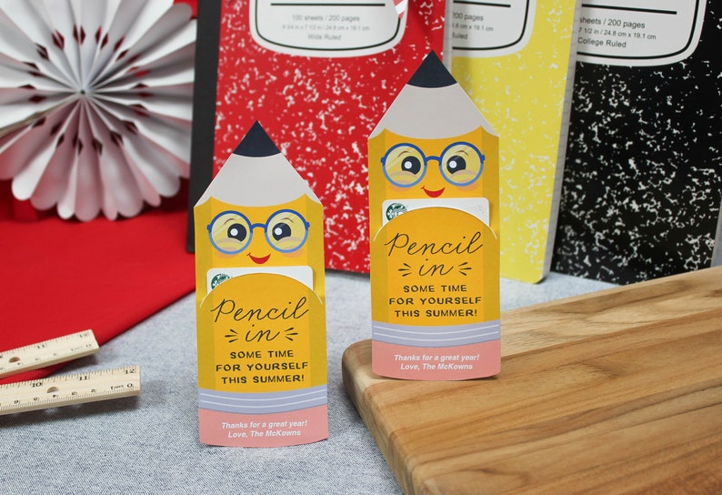 May include: Two yellow pencil-shaped gift card holders with smiling faces and glasses, with the text "Pencil in some time for yourself this summer!". The background includes composition notebooks and a decorative paper fan.