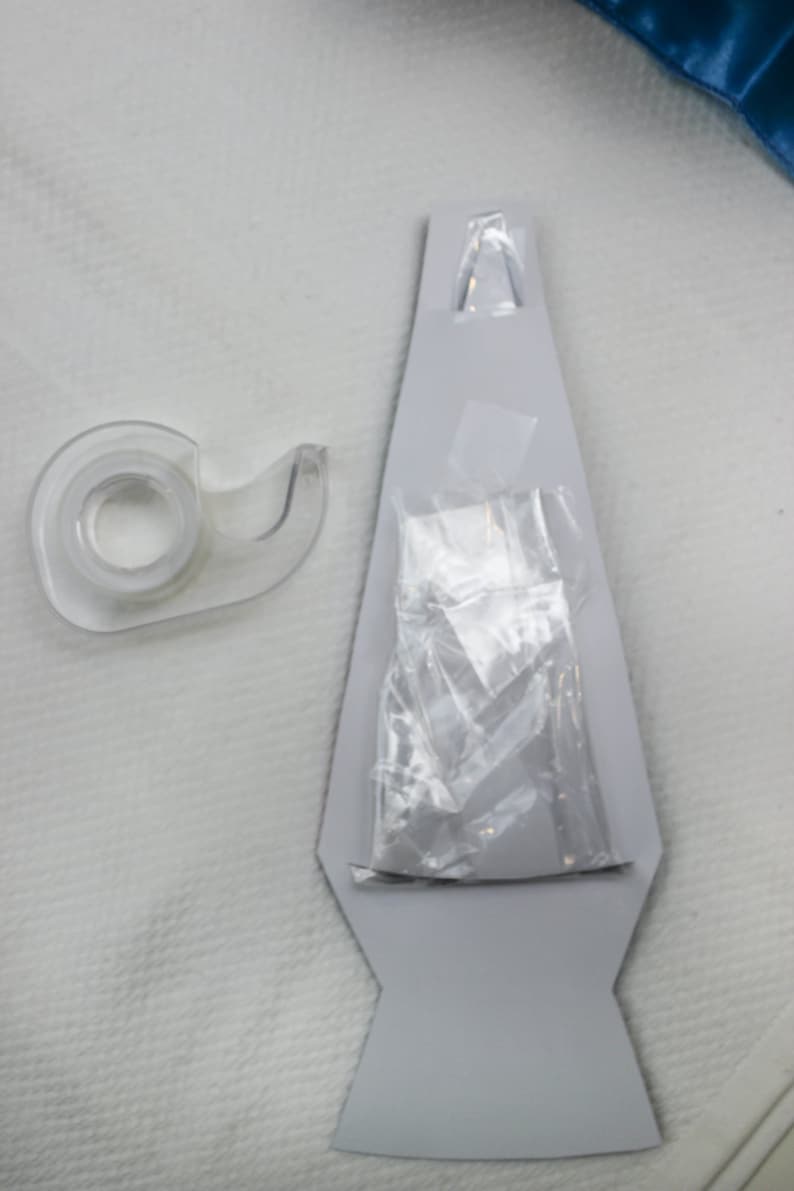 Peut inclure: Un distributeur de ruban adh&eacute;sif en plastique transparent sur fond blanc. Le ruban est partiellement d&eacute;roul&eacute; et il y a un sac en plastique transparent avec une &eacute;tiquette blanche &agrave; l'int&eacute;rieur d'une d&eacute;coupe en carton gris d'une lampe &agrave; lave.