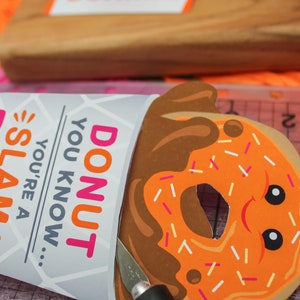 May include: A craft project featuring a donut-shaped paper cutout with chocolate icing and sprinkles. The donut has a hole in the center and googly eyes. The text "DONUT YOU KNOW... YOU'RE A SLAM" is printed on a white paper cup.