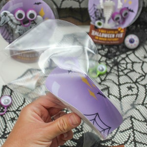 May include: A clear plastic bag with a purple and white Halloween-themed card inside. The card has a spiderweb design and the text "I see lots of Halloween fun in your future!" The bag is being held by a hand.