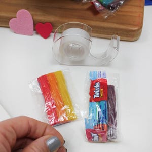 May include: A clear tape dispenser, two small packages of candy, and two felt hearts are on a white surface. One candy package contains rainbow-colored sticks, and the other contains Twizzlers. A wooden cutting board is in the background.