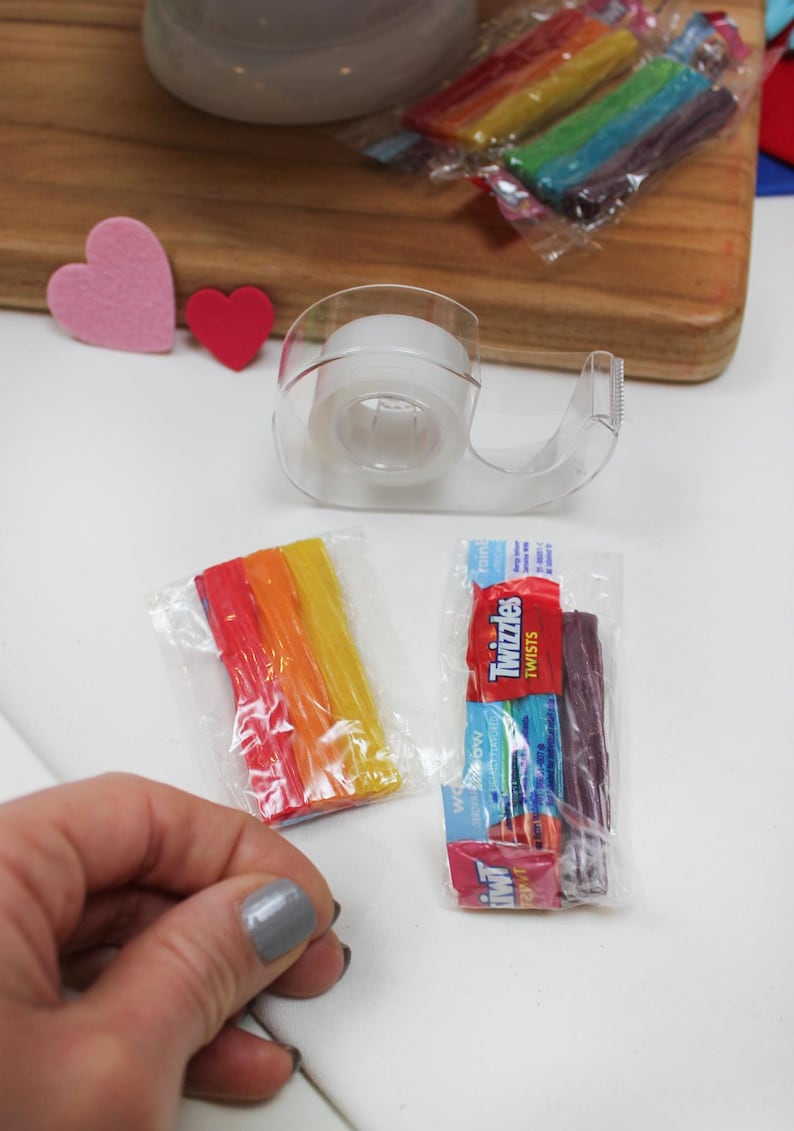 May include: A clear tape dispenser, two small packages of candy, and two felt hearts are on a white surface. One candy package contains rainbow-colored sticks, and the other contains Twizzlers. A wooden cutting board is in the background.