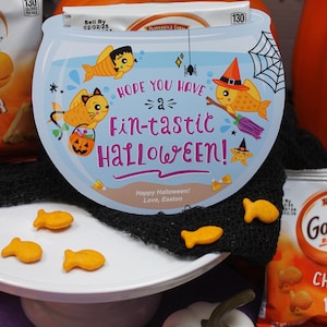 May include: A blue and white Halloween card with a fishbowl design. The card says "Hope you have a fin-tastic Halloween!" with a cartoon fish, a spider, and a pumpkin. There are also goldfish crackers on a white cake stand and a white pumpkin.