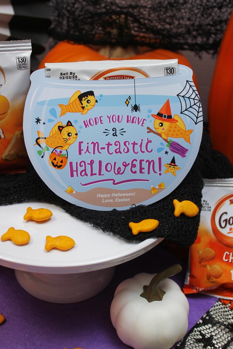May include: A blue and white Halloween card with a fishbowl design. The card says "Hope you have a fin-tastic Halloween!" with a cartoon fish, a spider, and a pumpkin. There are also goldfish crackers on a white cake stand and a white pumpkin.