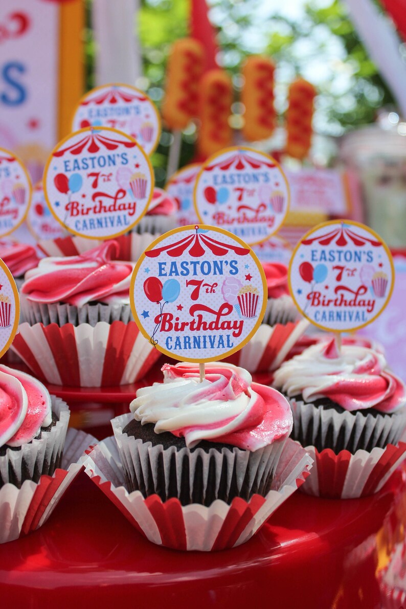 May include: A close-up of a table with cupcakes decorated with red and white frosting. Each cupcake has a round, colorful paper topper that says "Easton's 7th Birthday Carnival".