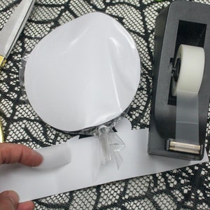 May include: A white circle sticker on a clear plastic sheet, a black and white patterned paper, a black tape dispenser with a white tape roll, and a pair of googly eyes on a green wire.