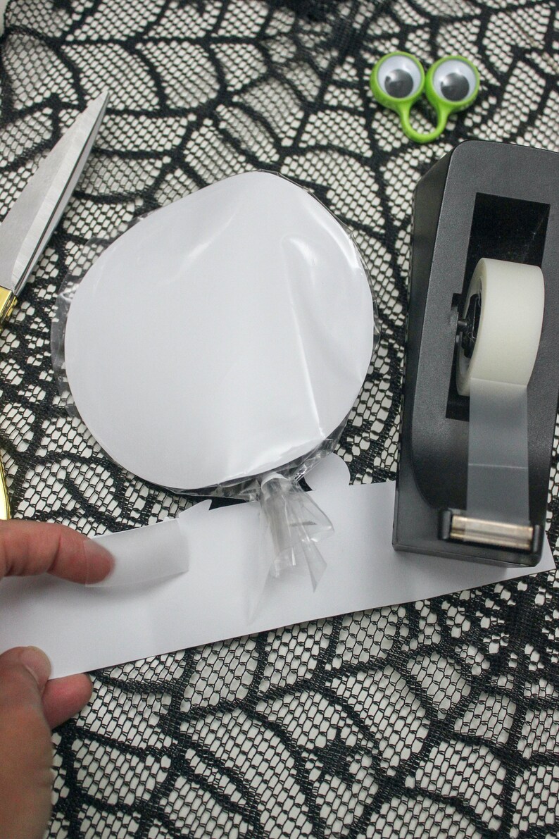 May include: A white circle sticker on a clear plastic sheet, a black and white patterned paper, a black tape dispenser with a white tape roll, and a pair of googly eyes on a green wire.