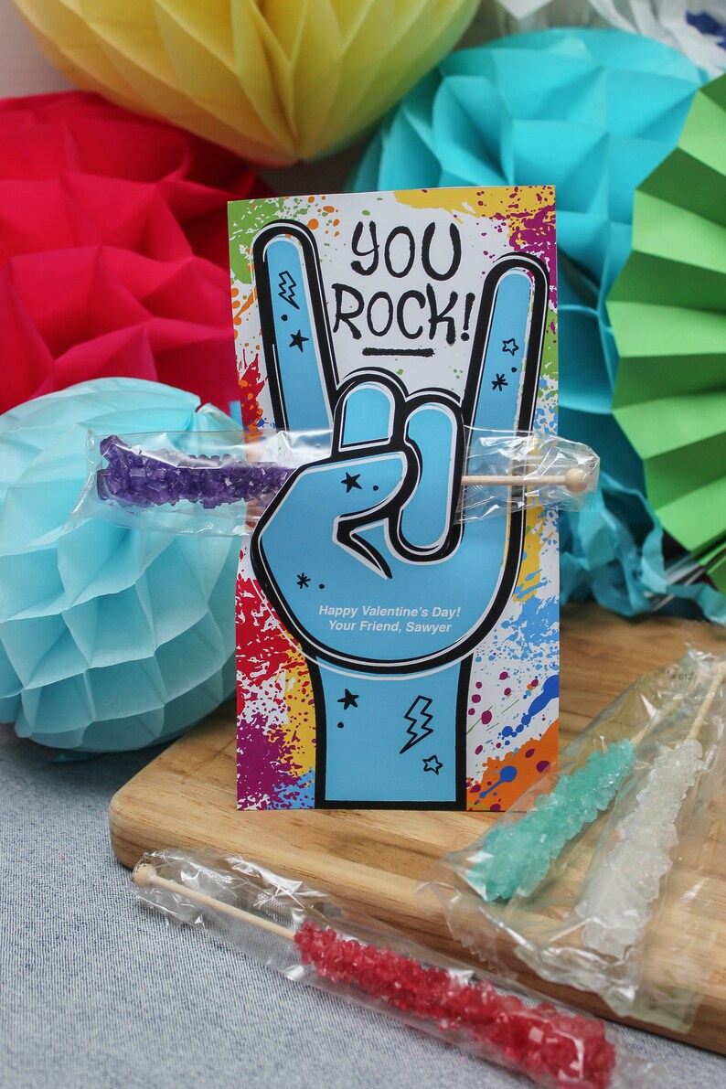 Pode incluir: Um cart&atilde;o colorido com uma m&atilde;o azul fazendo o sinal de rock and roll. O cart&atilde;o diz "You Rock!" e "Happy Valentine's Day! Your Friend, Sawyer". O cart&atilde;o est&aacute; rodeado de papel de seda colorido e pirulitos de a&ccedil;&uacute;car.