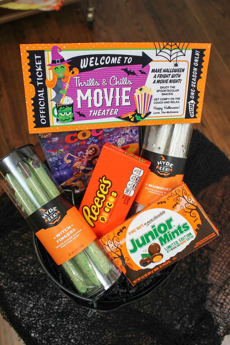 May include: A black bucket filled with Halloween-themed snacks, including a movie ticket, Reese's Pieces, Junior Mints, and pretzel rods. The ticket reads "Welcome to Thrills & Chills Movie Theater".