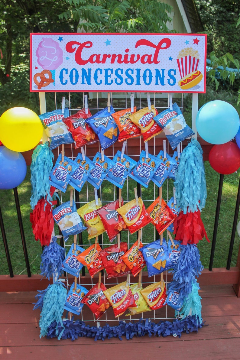 May include: A white wire rack with various bags of chips hanging from it. The bags are all different brands and flavors, including Ruffles, Doritos, Cheetos, and Fritos. The rack is decorated with blue, red, and white streamers. A sign above the rack reads "Carnival Concessions".