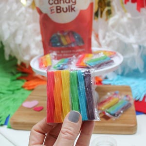 May include: A hand holds a package of rainbow-colored candy sticks. The candy is arranged in a spectrum of colors, from red to purple. A bag of "Candy in Bulk" is in the background, along with other candies on a white plate and a wooden cutting board.
