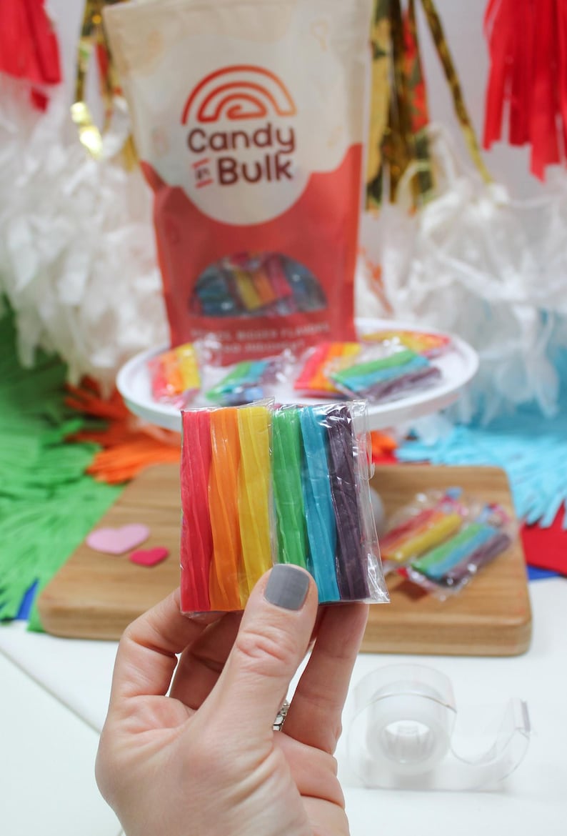 May include: A hand holds a package of rainbow-colored candy sticks. The candy is arranged in a spectrum of colors, from red to purple. A bag of "Candy in Bulk" is in the background, along with other candies on a white plate and a wooden cutting board.