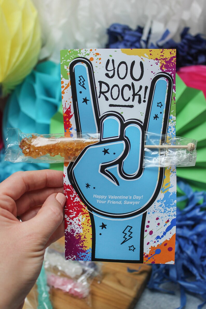 Pode incluir: Um cart&atilde;o colorido com uma m&atilde;o azul fazendo o sinal de rock and roll. O cart&atilde;o diz "You Rock!" e "Happy Valentine's Day! Your Friend, Sawyer". Um pirulito de rocha marrom est&aacute; preso ao cart&atilde;o.