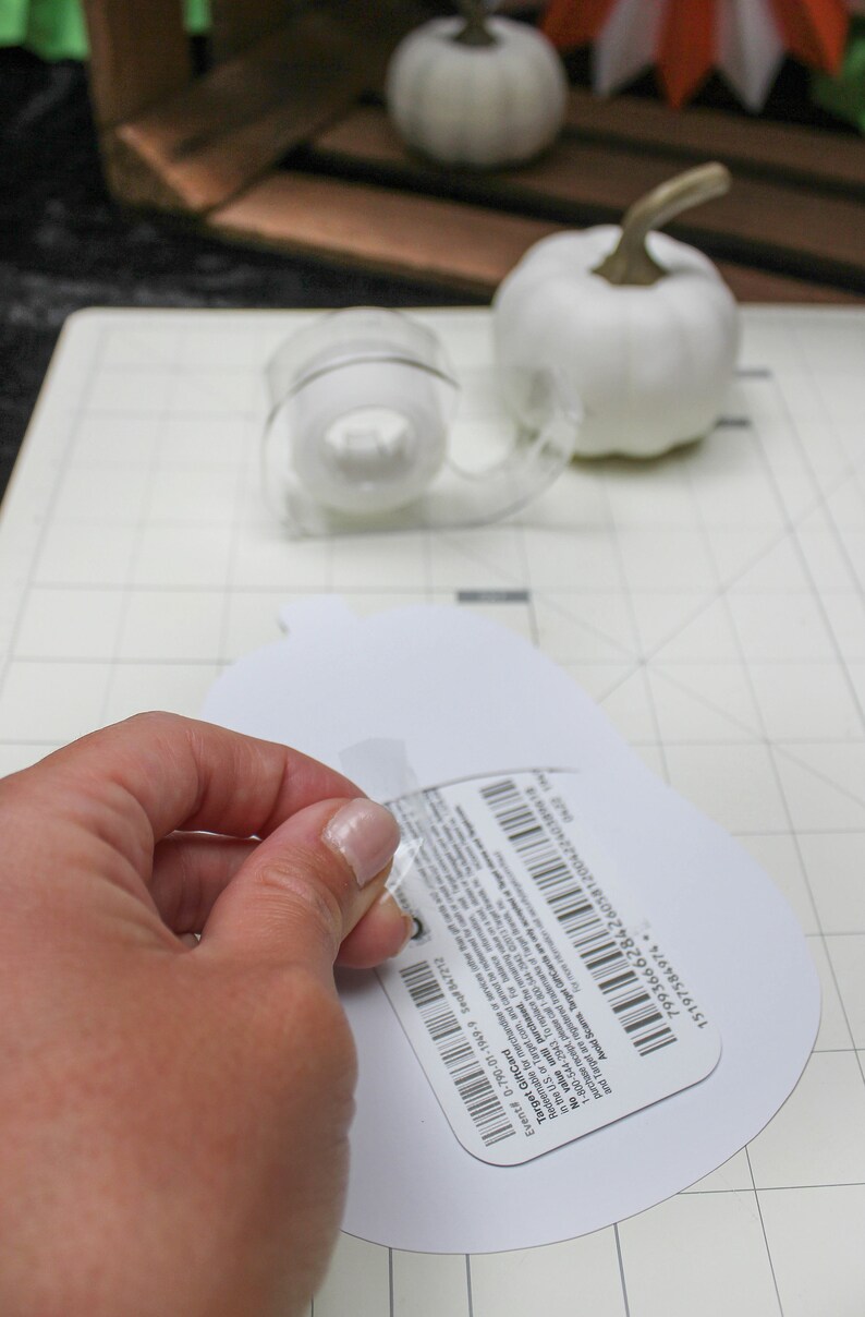 May include: A white pumpkin cutout with a Target gift card attached to it. The gift card has a barcode and the text "Target Gift Card" printed on it. The gift card is being peeled off of the pumpkin cutout.