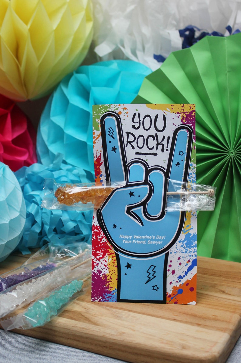 Pode incluir: Um cart&atilde;o colorido com as palavras "You Rock!" e uma m&atilde;o azul fazendo o gesto de rock and roll. O cart&atilde;o est&aacute; rodeado de decora&ccedil;&otilde;es de papel coloridas. O cart&atilde;o tem o texto "Happy Valentine's Day! Your Friend, Sawyer" na parte inferior.