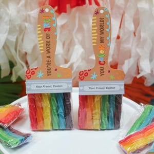 May include: Two paint brush-shaped gifts with rainbow-colored candy bristles. The wooden handles have the phrases "YOU'RE A WORK OF ART!" and "YOU Color MY WORLD!" with the name "Easton" printed on them. The gifts are on a white cake stand.