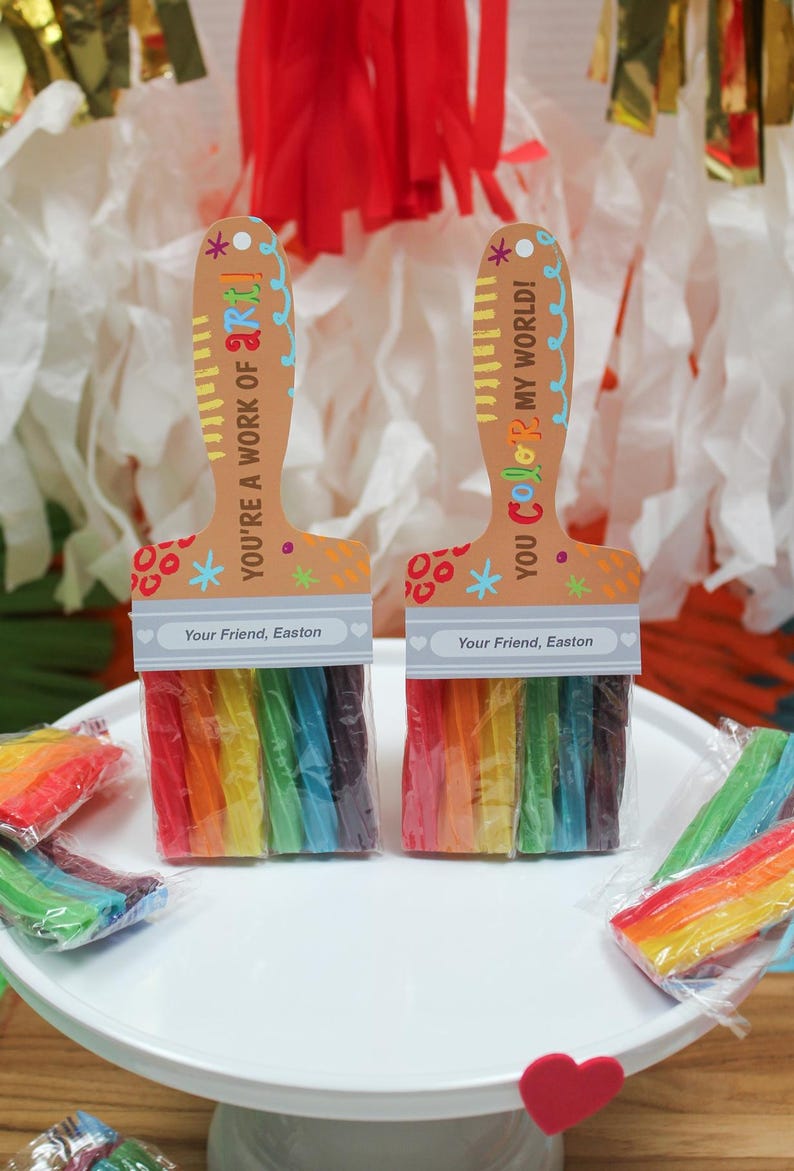 May include: Two paint brush-shaped gifts with rainbow-colored candy bristles. The wooden handles have the phrases "YOU'RE A WORK OF ART!" and "YOU Color MY WORLD!" with the name "Easton" printed on them. The gifts are on a white cake stand.