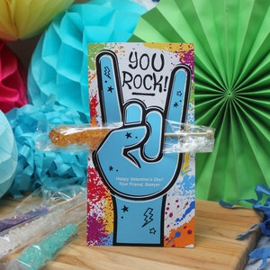 Pode incluir: Um cart&atilde;o colorido com o texto "You Rock!" e uma m&atilde;o azul fazendo o gesto de rock and roll. O cart&atilde;o est&aacute; apoiado em um saco de pl&aacute;stico transparente que cont&eacute;m um palito de caramelo de rocha.