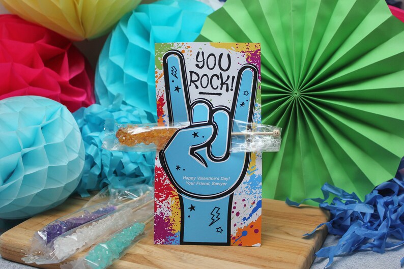 Pode incluir: Um cart&atilde;o colorido com o texto "You Rock!" e uma m&atilde;o azul fazendo o gesto de rock and roll. O cart&atilde;o est&aacute; apoiado em um saco de pl&aacute;stico transparente que cont&eacute;m um palito de caramelo de rocha.