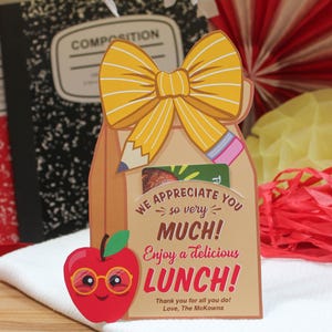 May include: A brown paper bag with a yellow bow, a red apple with googly eyes and glasses, and the text "WE APPRECIATE YOU so very MUCH! Enjoy a delicious LUNCH! Thank you for all you do! Love, The McKowns".