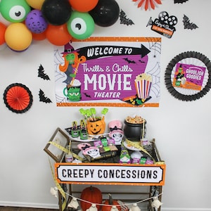 May include: A Halloween-themed movie night party setup with a gold bar cart decorated with a banner that says "Creepy Concessions". The cart is filled with various Halloween-themed treats, including a pumpkin-shaped bowl filled with green and white marshmallows, a cauldron filled with popcorn, and a variety of other snacks. The background features a purple and orange poster with the text "Welcome to Thrills & Chills Movie Theater" and a spiderweb design. There are also black and orange paper fans, black bats, and a garland of orange, yellow, and green balloons with black eyes.