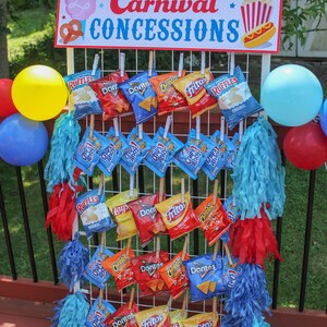 May include: A white wire rack with various bags of chips hanging from it. The bags are all different brands and flavors, including Ruffles, Lay's, Doritos, Cheetos, and Fritos. The rack is decorated with blue and red streamers. A sign above the rack reads "Carnival Concessions".