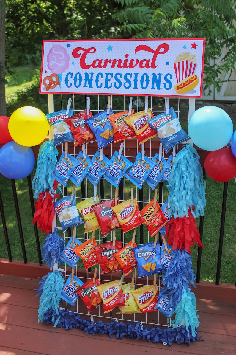May include: A white wire rack with various bags of chips hanging from it. The bags are all different brands and flavors, including Ruffles, Lay's, Doritos, Cheetos, and Fritos. The rack is decorated with blue and red streamers. A sign above the rack reads "Carnival Concessions".