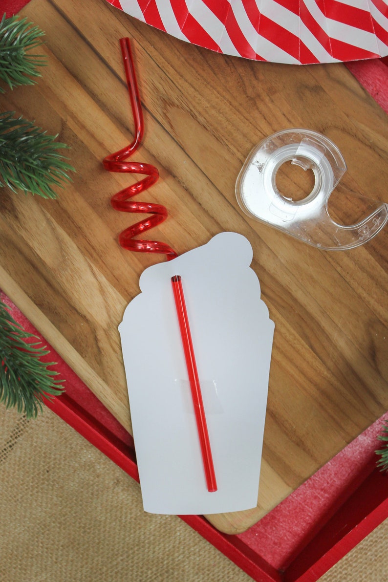May include: A white paper cutout of a coffee cup with a red straw sticking out of the top. The straw is curled into a spiral shape. The cup is sitting on a wooden surface.