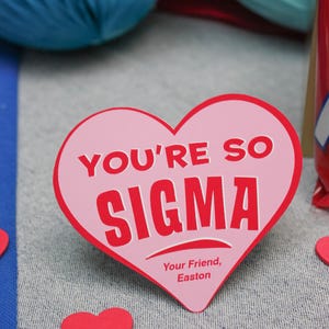 May include: Pink heart-shaped card with the text "You're so Sigma" in red letters. The card also has the text "Your Friend, Easton" in smaller red letters.
