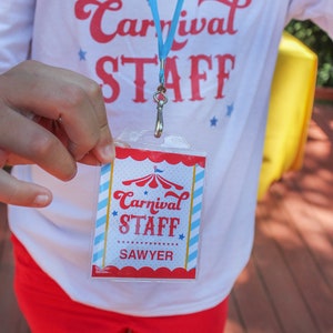 May include: A white t-shirt with red text that says "Carnival Staff" and a blue lanyard with a red, white, and blue carnival tent design. A clear plastic badge with red, white, and blue stripes and a carnival tent design says "Carnival Staff Sawyer".