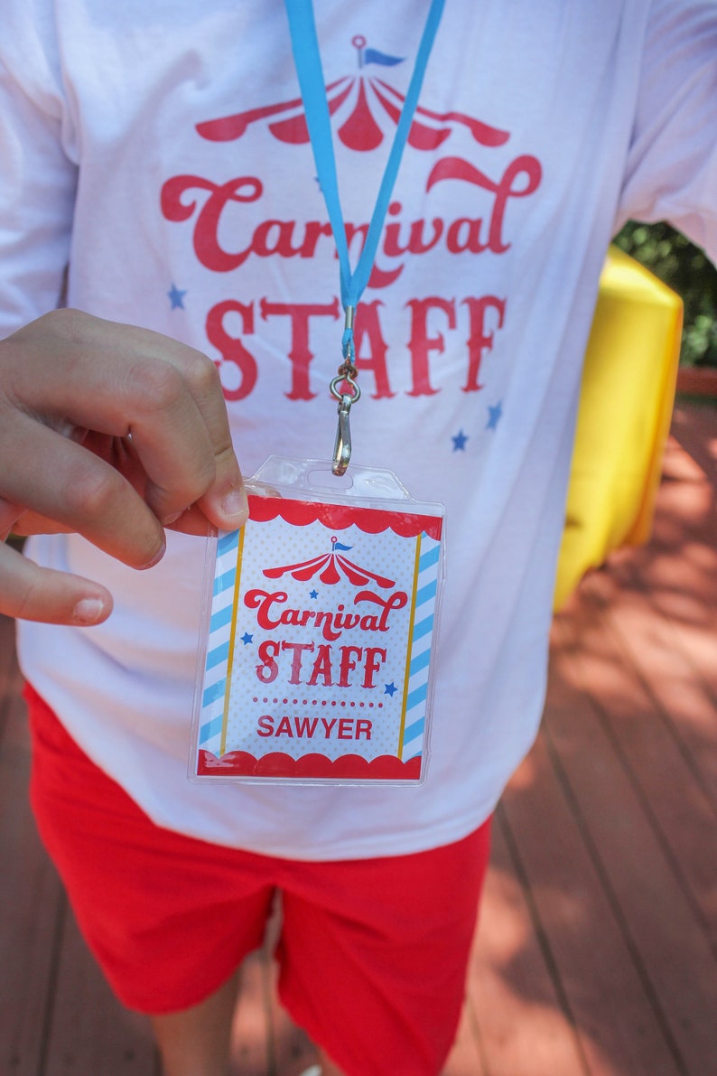 May include: A white t-shirt with red text that says "Carnival Staff" and a blue lanyard with a red, white, and blue carnival tent design. A clear plastic badge with red, white, and blue stripes and a carnival tent design says "Carnival Staff Sawyer".
