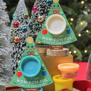 May include: Two Play-Doh containers shaped like Christmas trees with a Play-Doh container inside each. The trees are green with a white snowflake design and a red bow. The containers are labeled "It's the most Won-Doh-Ful Time of the Year!" and "Your Friend, Easton".