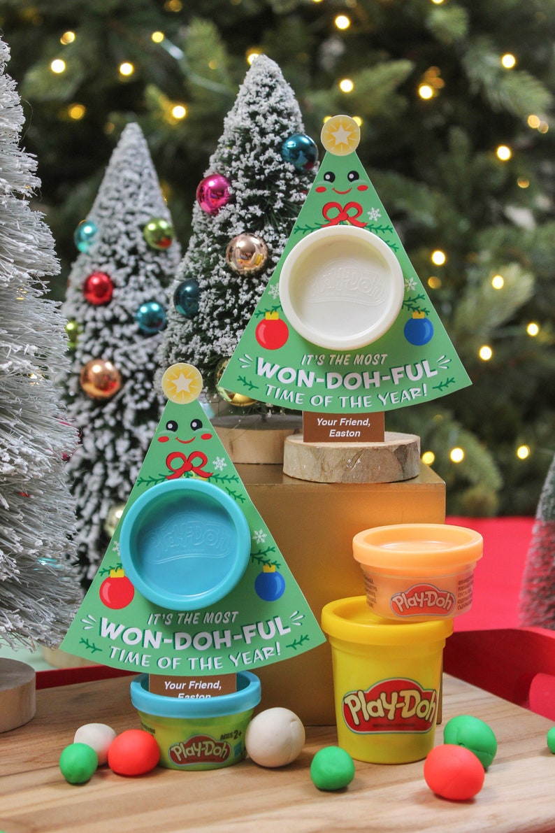 May include: Two Play-Doh containers shaped like Christmas trees with a Play-Doh container inside each. The trees are green with a white snowflake design and a red bow. The containers are labeled "It's the most Won-Doh-Ful Time of the Year!" and "Your Friend, Easton".
