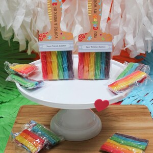 May include: Two paint brush-shaped gifts with rainbow-colored candy. The paint brushes have a wooden handle design with the text "YOU'RE A WORK OF ART!" and "YOU COLOR MY WORLD!" The gifts are on a white cake stand and wooden cutting board.