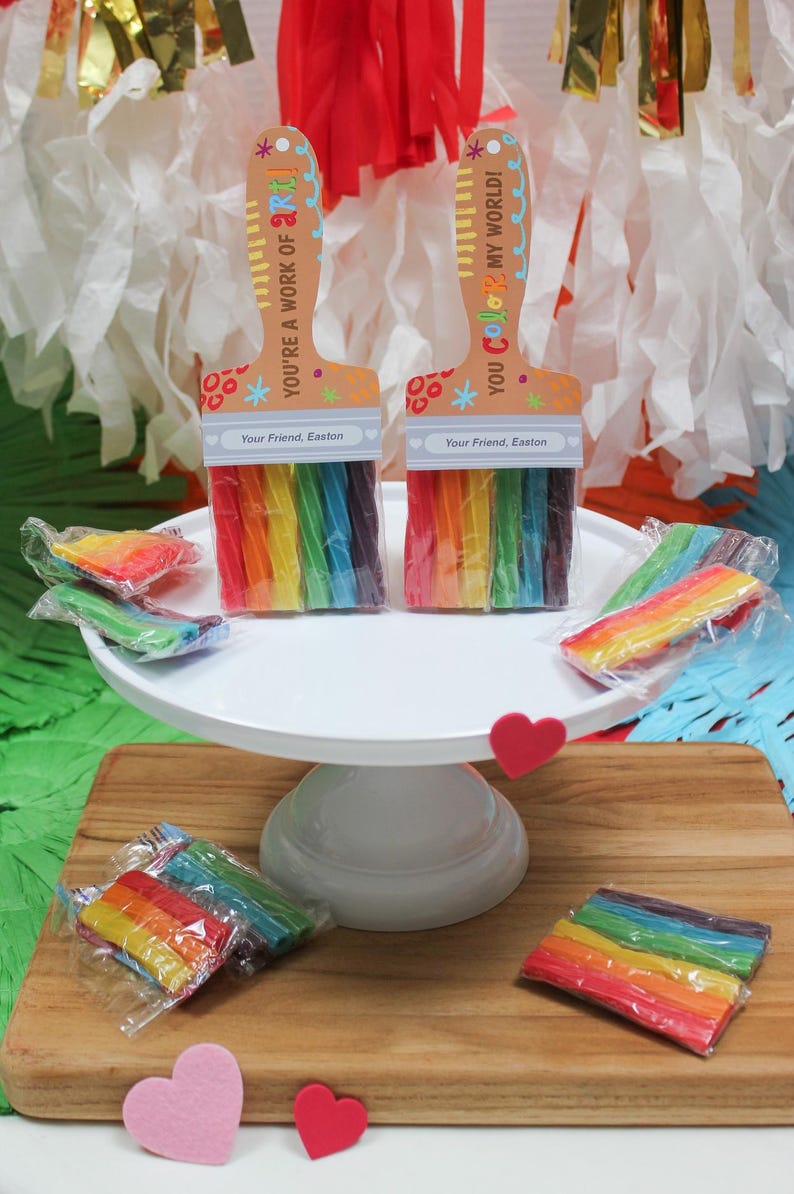 May include: Two paint brush-shaped gifts with rainbow-colored candy. The paint brushes have a wooden handle design with the text "YOU'RE A WORK OF ART!" and "YOU COLOR MY WORLD!" The gifts are on a white cake stand and wooden cutting board.