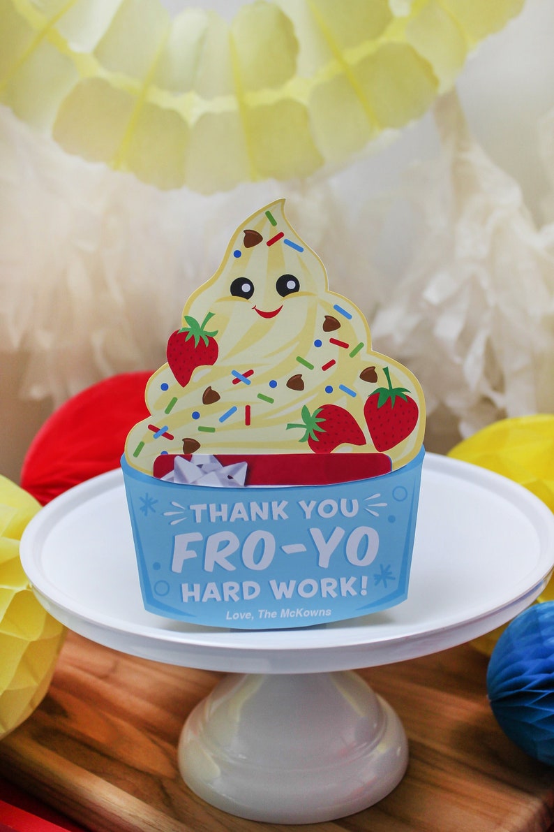May include: A white cake stand with a blue and white card on top. The card features a cartoon yogurt cup with a smiling face and the text "Thank you Fro-Yo Hard Work! Love, The McKowns".
